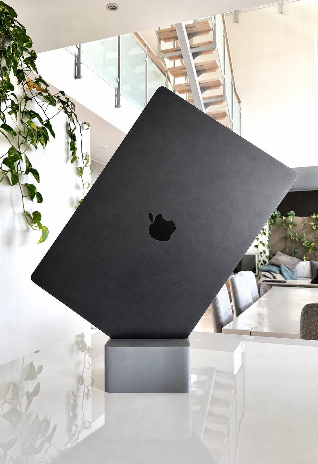 A Cubistand holding a closed black MacBook Pro upright on a glass table, with houseplants and a staircase behind it